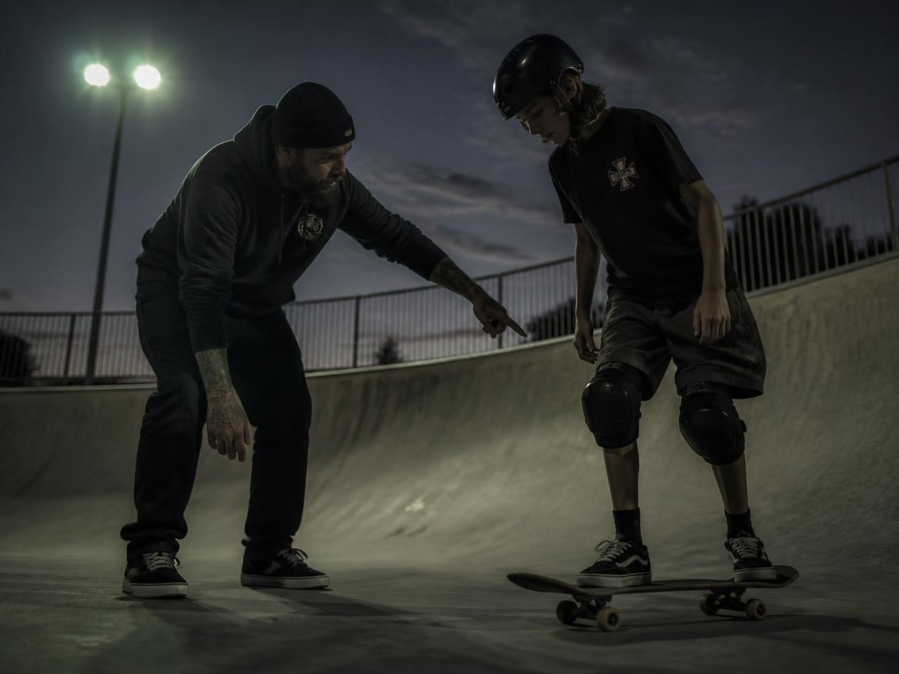 A skate coach guiding a young rider on the edge of a concrete bowl at dusk